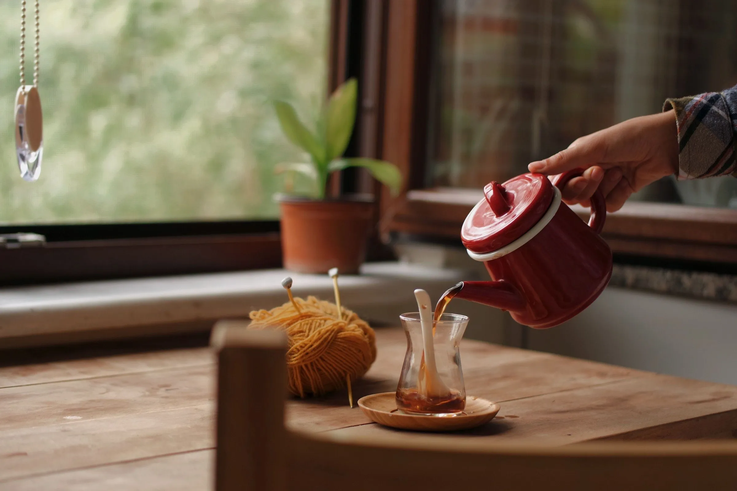 A hand pouring tea from a red enamel pot into a glass on a wooden kitchen table by the window