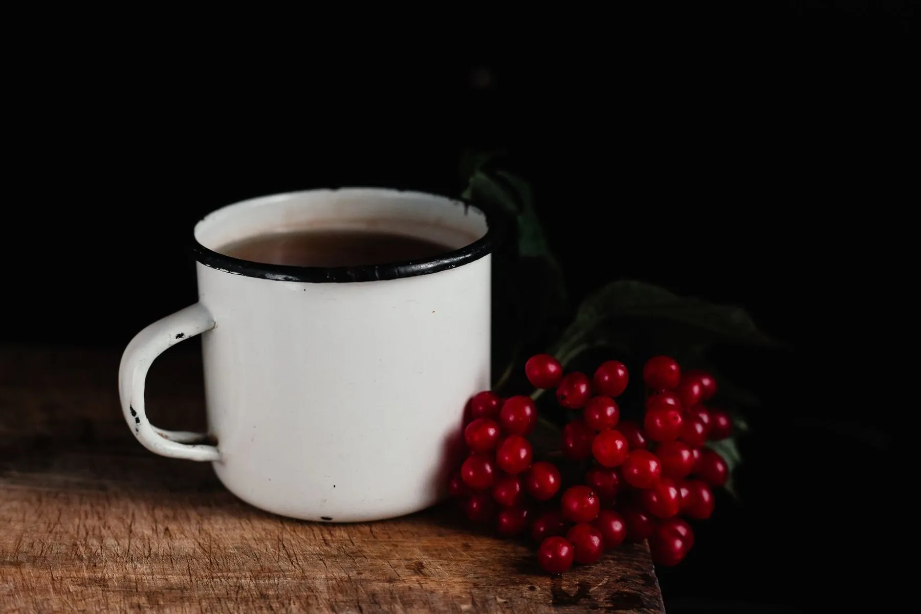 A well-used white enamel mug with a chipped handle beside red berries on dark wood