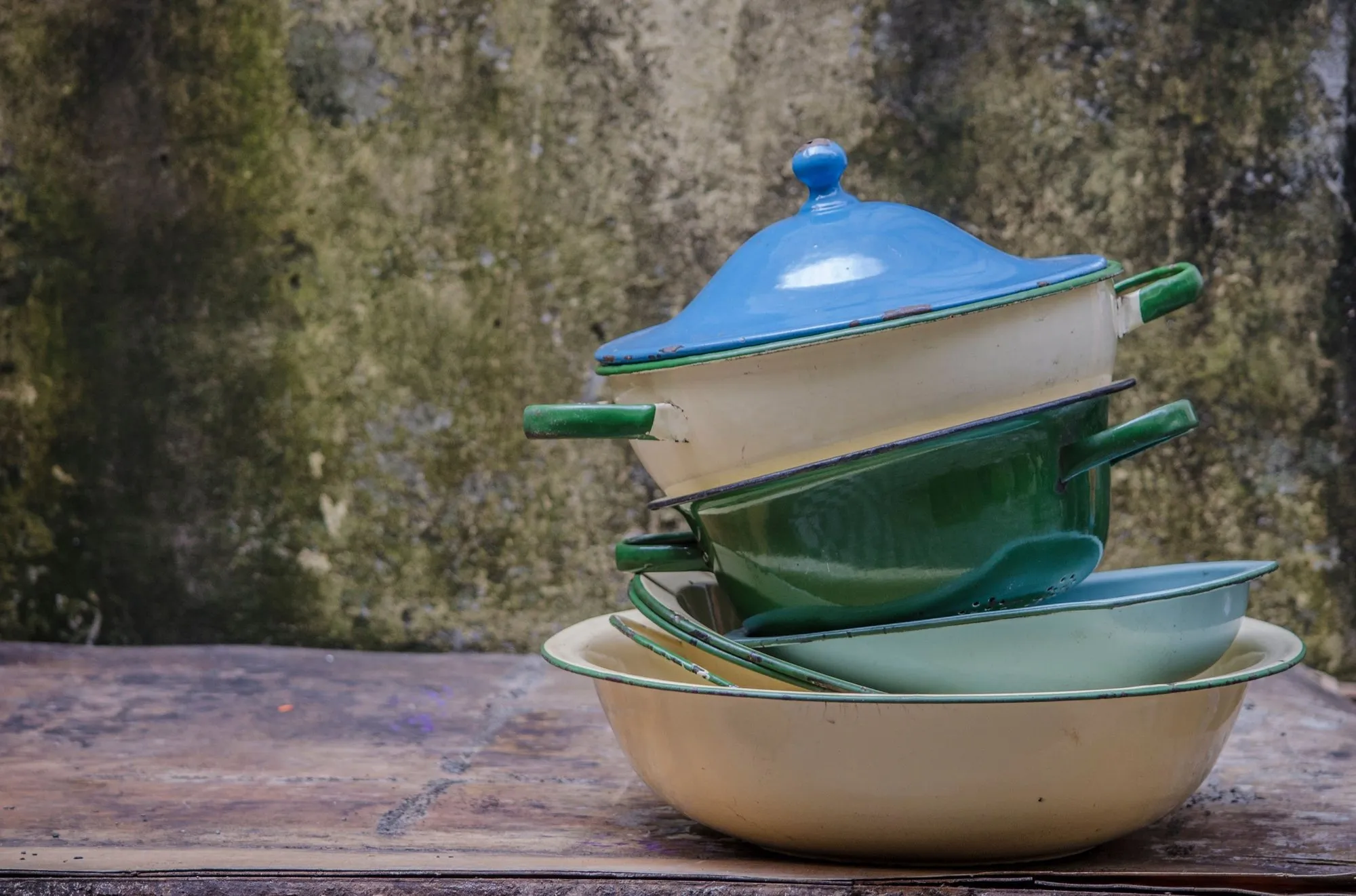 A stack of cream, green and blue enamel bowls and pots on a weathered wooden table